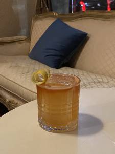 Fitzgerald Em um copo redondo sobre uma mesa, é servido drinque de cor alaranjada decorado com casca de laranja