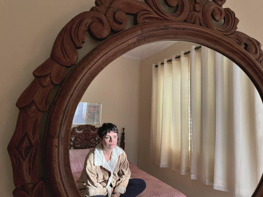 Renata Gaspar Mulher está sentada em uma cama e dá para vê-la pelo reflexo de um espelho