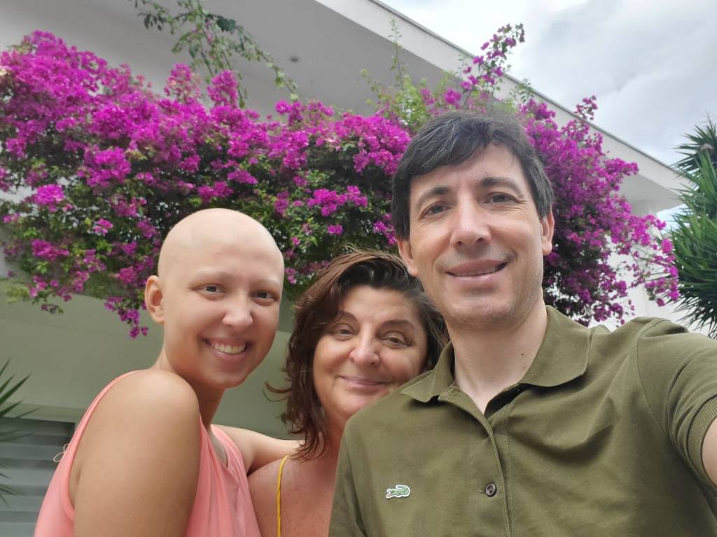 – Victoria (filha), Lourdes Edreira e Victor Angueira (ARQUIVO PESSOAL) A filha Victoria ao lado dos pais Lourdes e Victor. Os três aparecem lado a lado em frente a uma plantação de flores roxas.
