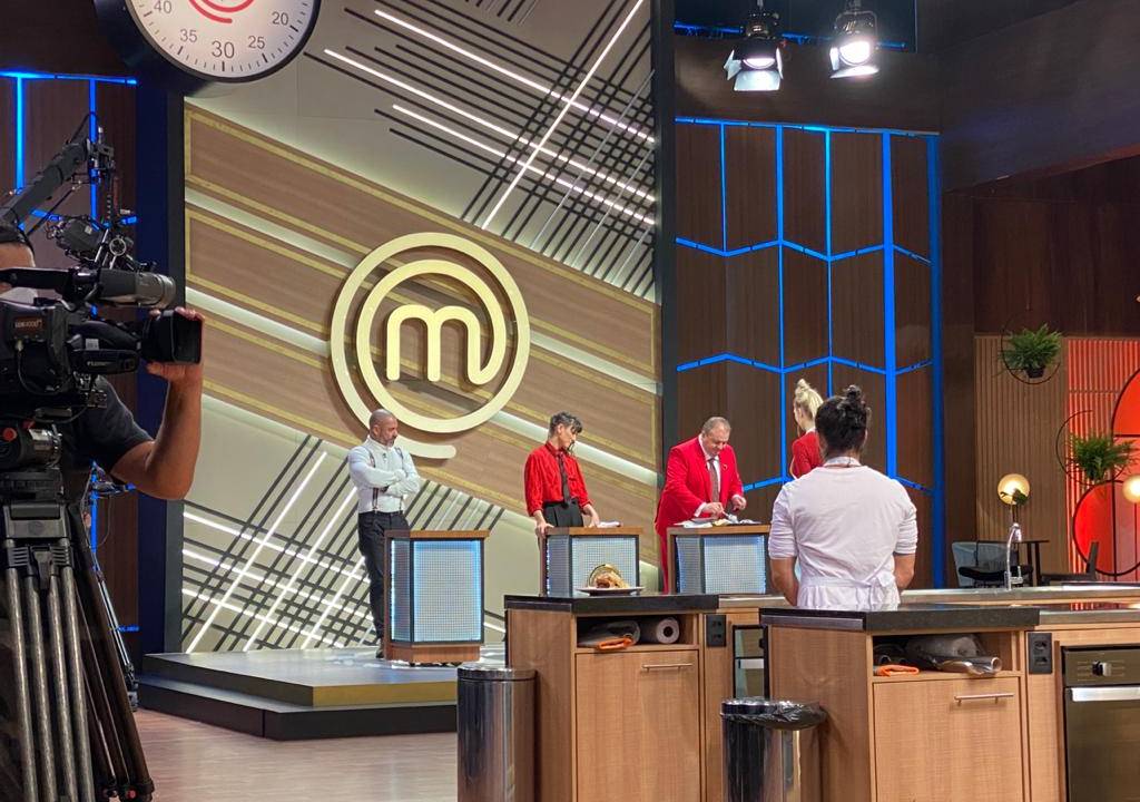 Set do programa MasterChef Brasil, com jurados no tablado ao fundo e participantes à esquerda.