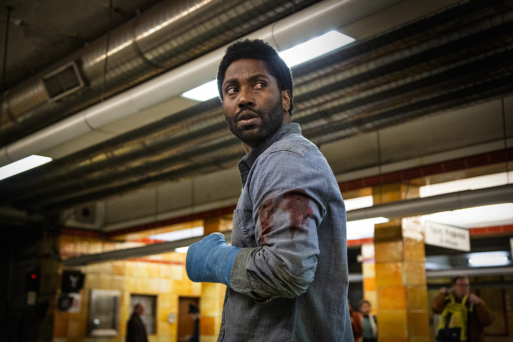 A imagem mostra John David Washington atuando em Beckett. A câmera mostra ele da cintura para cima e seu casaco está com marcas de sangue na altura do tríceps.