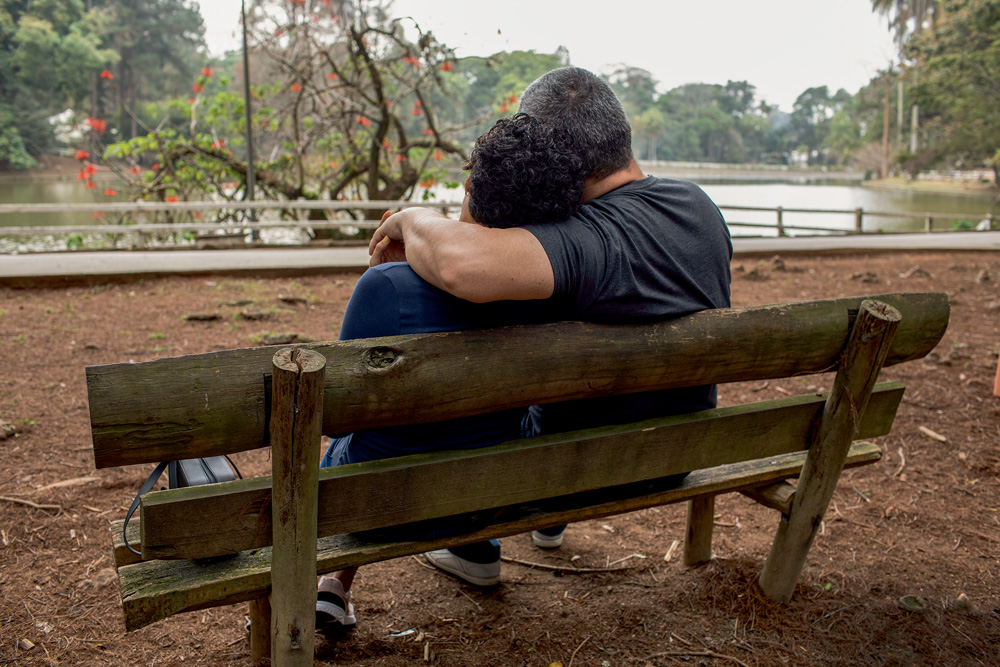 beatriz* e otávio* sentados em um banco de parque olhando para frente; otávio* está com seu braço sobre os ombros de beatriz*