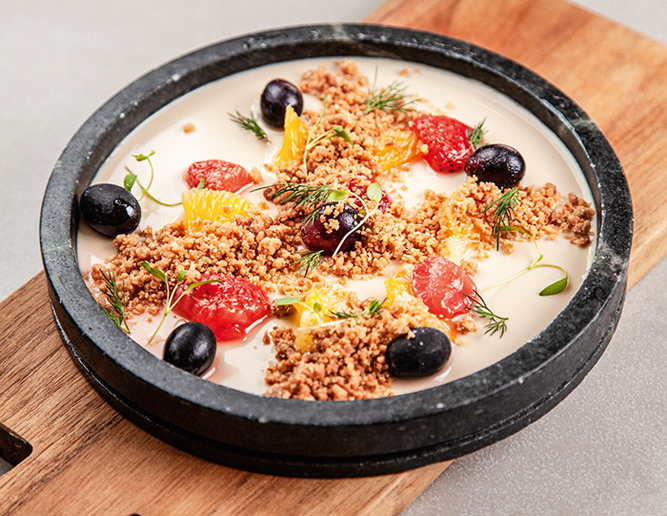 Davo Cozinha Sobre uma tábua de madeira, está um prato raso de pedra com panacota coberta por crumble de sucrilhos, doce de morango e gelo de uva.