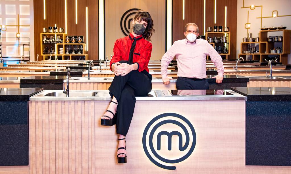 Helena Rizzo e Arnaldo Lorençato A chef Helena Rizzo sentada na bancada do MasterChef e Arnaldo Lorençato encostado na bancada logo atrás, à direita. Ambos de máscara.