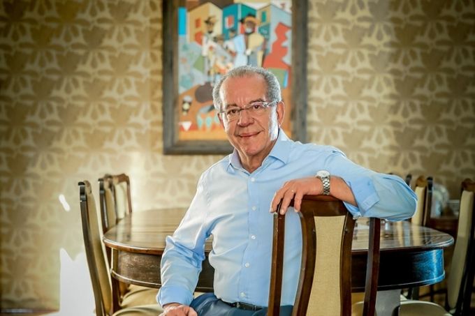 josé aníbal sentado em cadeira em sua sala de jantar. ele está de lado na cadeira, com o braço esquerdo apoiado no suporte da cadeira. ele está sorrindo, usando óculos e camisa azul. na parede ao fundo, há papel de parede beje escuro com detalhes em branco e um quadro colorido