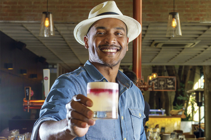 Em frente ao bar, Laércio Zulu posa vestindo uma camisa azul e um chapéu branco, segurando um copo de drinque.