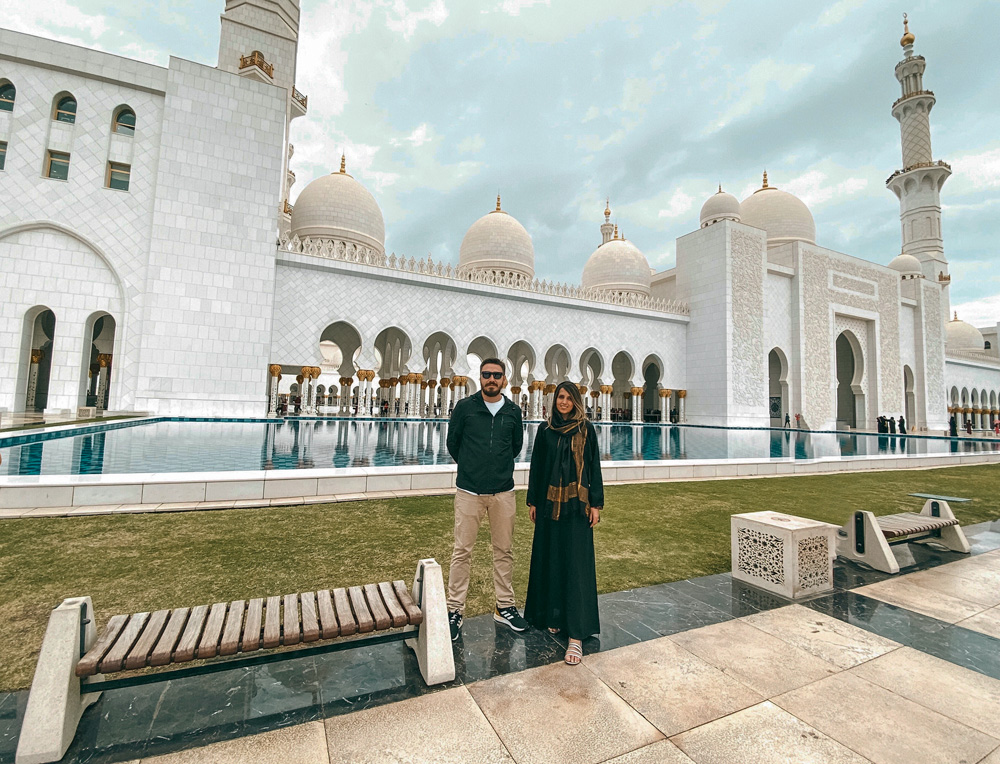 mesquita-abu-dhabi-lua-de-mel-tati-gabriel-casal-viajante-nosso-louco-amor tati e gabriel posando para a foto com mesquita ao fundo, em abu dhabi