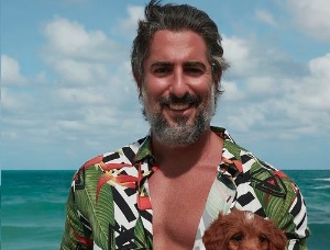 Foto mostra Mion em praia, com camisa aberta, mostrando parcialmente o peito. Ele sorri