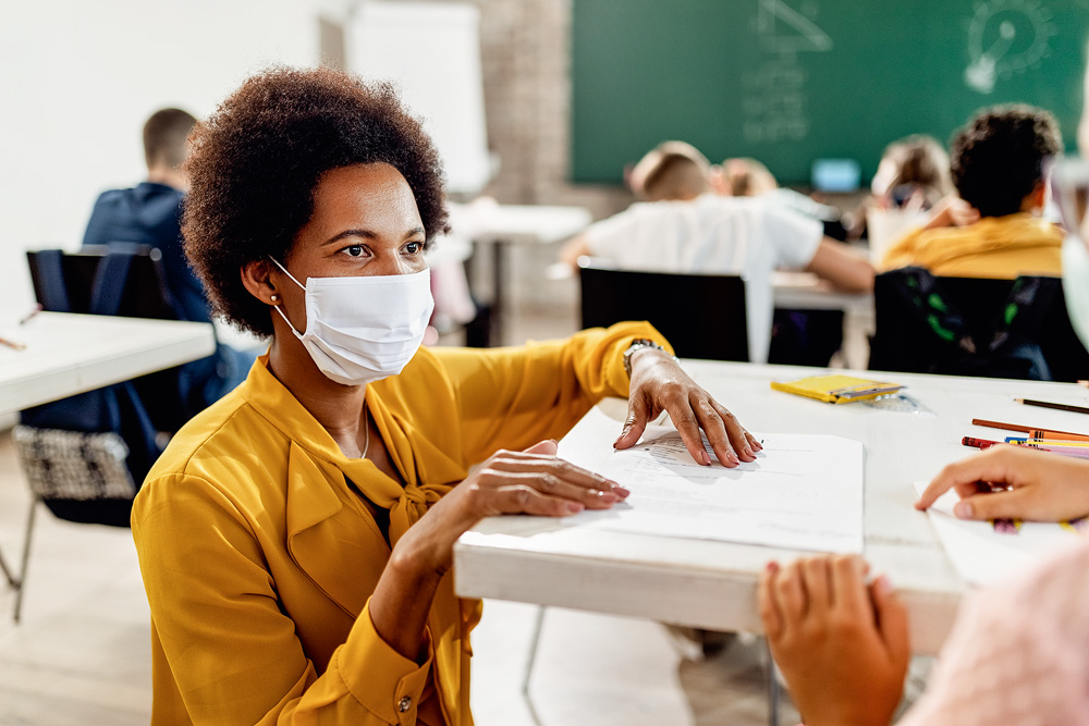 professora negra de máscara em uma sala de aula com as mãos na mesa de um aluno
