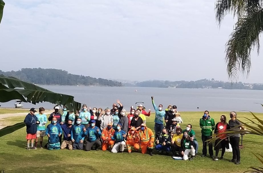 Um grupo de pessoas de várias idades posa em frente a Represa de Guarapiranga; dia está nublado