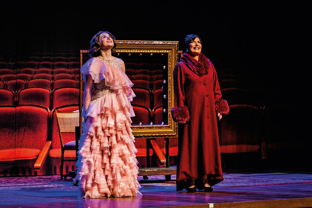retorno-teatros-peça-som-e-sílaba-Alessandra-Maestrini-Mirna-Rubim-olhando-para-cima-contracenando.jpg alessandra maestrini e mirna rubim uma ao lado da outra contracenando. elas estão de pé, olhando para cima. alessandra está com um vestido exuberante rendado rosa e mirna usa espécie de roupão vermelho