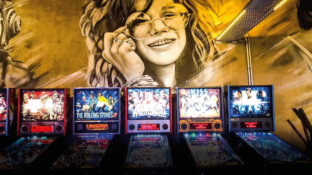 Parede do bar Bario decorada com gravura da Janis Joplin. Junto da parede, aparelhos de fliperama.