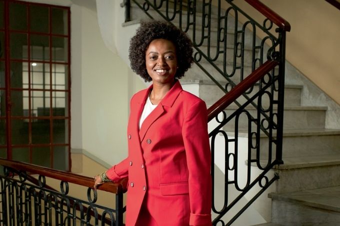 aline torres de terno vermelho sorrindo para a foto nas escadas do edifício da secretaria de cultura. ela está se apoiando com a mão direita no corrimão da escada