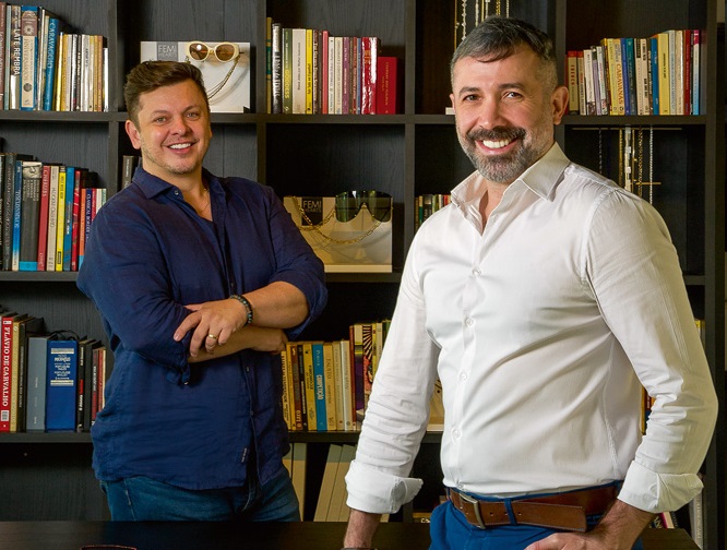 Dois homens em uma biblioteca posam para a foto utilizando roupas sociais