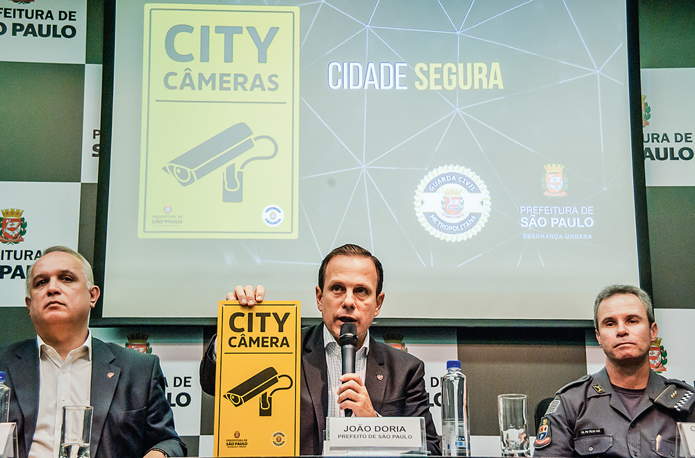 Doria-anuncia-projeto-city-câmeras-fracasso-projeto-baixa-adesão-público-prefeitura-promete-repaginação joão doria sentado discursando fazendo anúncio do City câmeras, segurando placa do projeto e com slide projetado atrás dele