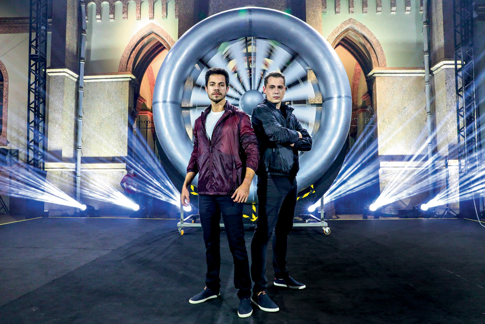 Henry e Klauss posando para a foto um ao lado do outro, meio de costas um para o outro, em cenário do novo programa. klaus está de braços cruzados