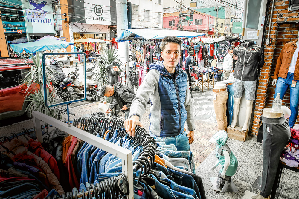 LAUROPIMENTA_1.JPG A imagem mostra Pimenta, com uma mão sob uma estrutura de cabides de roupa e olhando para a câmera na Rua Oriente.