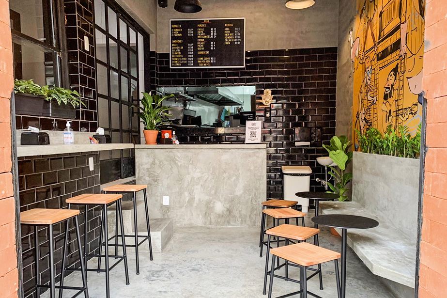 Parede de azulejos pretos decoram a lanchonete junto de banquetas com assento de madeira, mesas metálicas de cor preta, um quadro com as opções do menu e algumas plantas espalhadas pelo ambiente