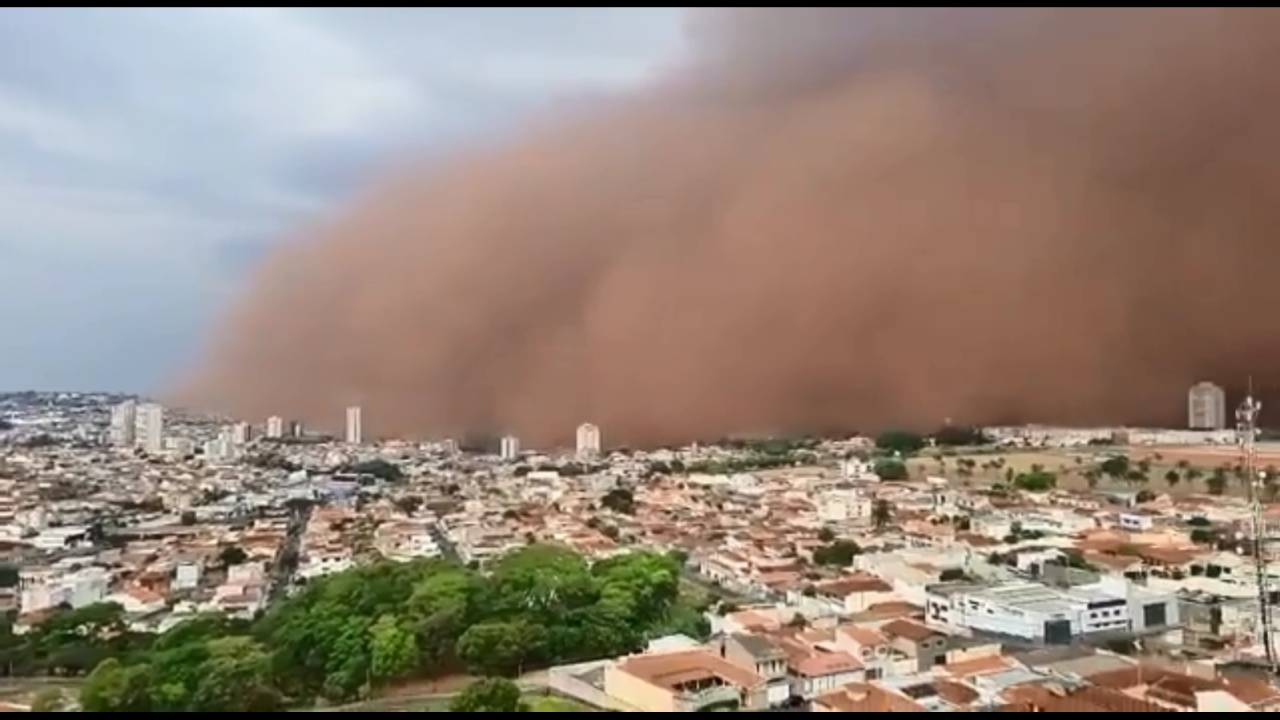 A imagem mostra uma foto ampla de uma cidade e, ao fundo, uma grande nuvem de poeiro encobre parte dela.