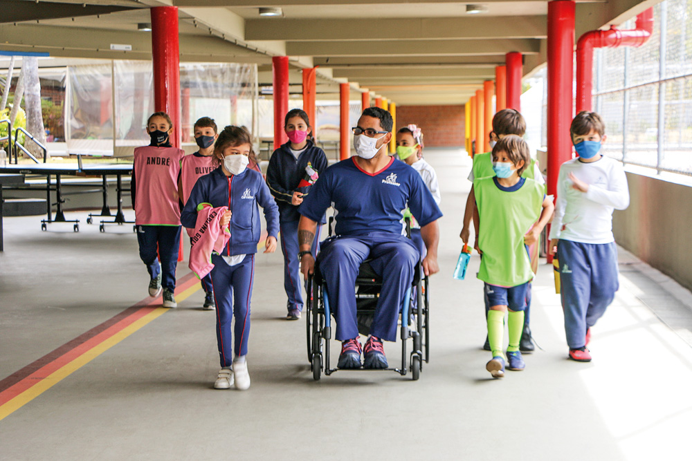André de Almeida Marques em cadeira de rodas andando por corredor semiaberto da escola Miguel de Cervantes com alunos pequenos ao redor. Todos estão de máscara