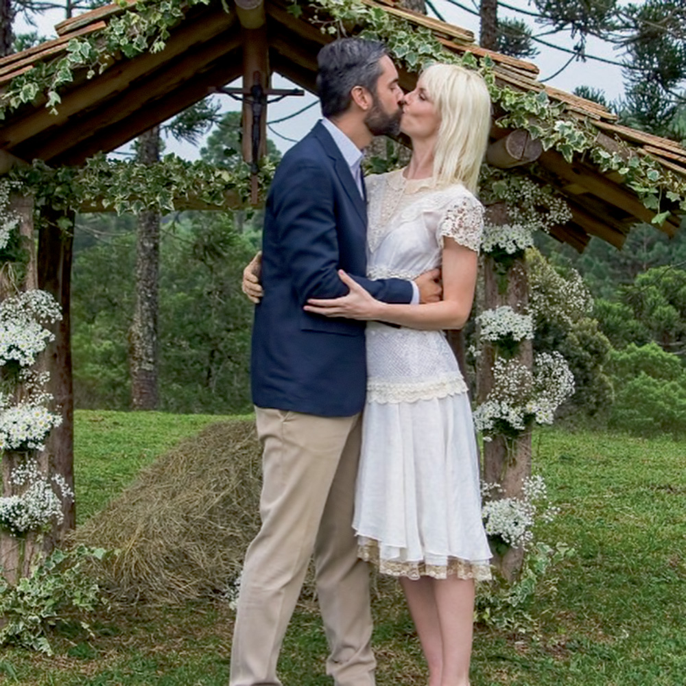 augusto-botelho-ana-claudia-michels-casamento-quase-afundou-barco-Nosso-louco-amor augusto e ana se beijando, ele com um terno, ela de vestido branco, em seu casamento, em um local aberto com gramado e diversas árvores. atrás deles há uma estrutura estilo portal de madeira ornada com flores
