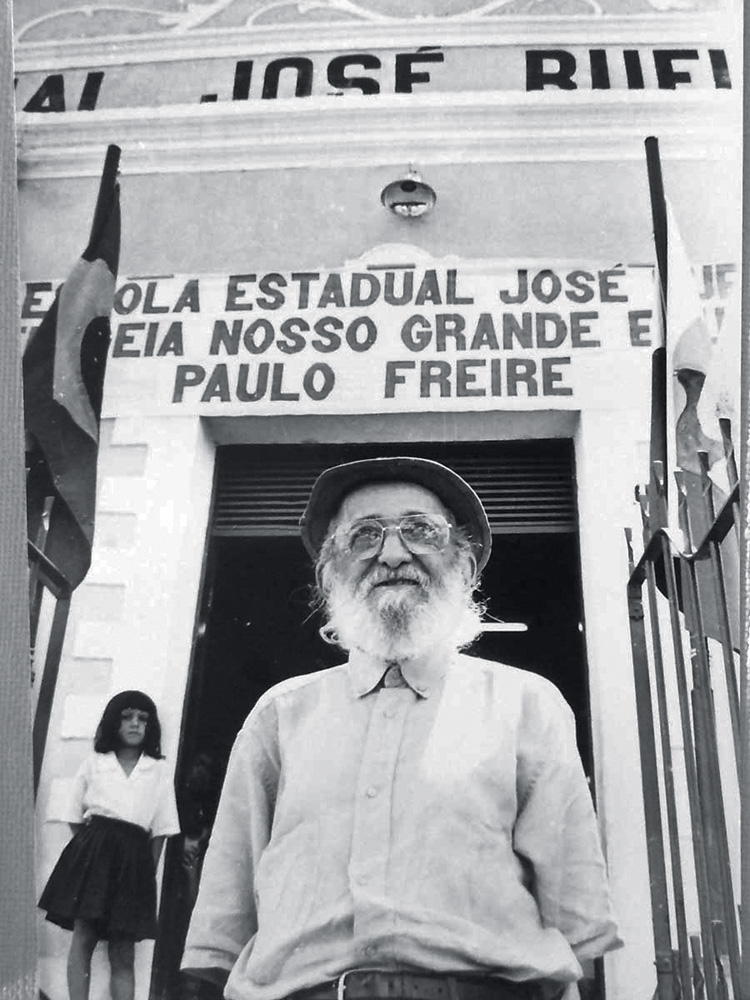 foto-Paulo-Freire-exposição-centenário-Paulo-Freire foto em preto e branco de paulo freire, que está olhando para o horizonte com barba longa branca e chapéu em frente a uma escola estadual que leva seu nome