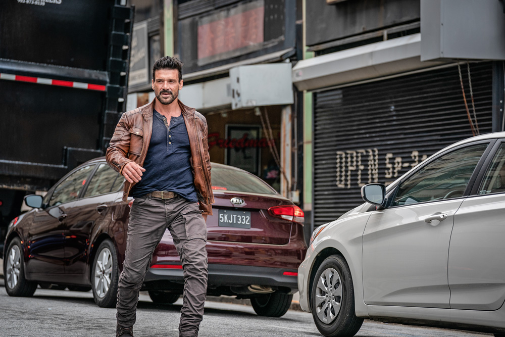 A imagem mostra Frank Grillo, com calça jeans e jaqueta de couro, andando no meio de uma rua de uma cidade.