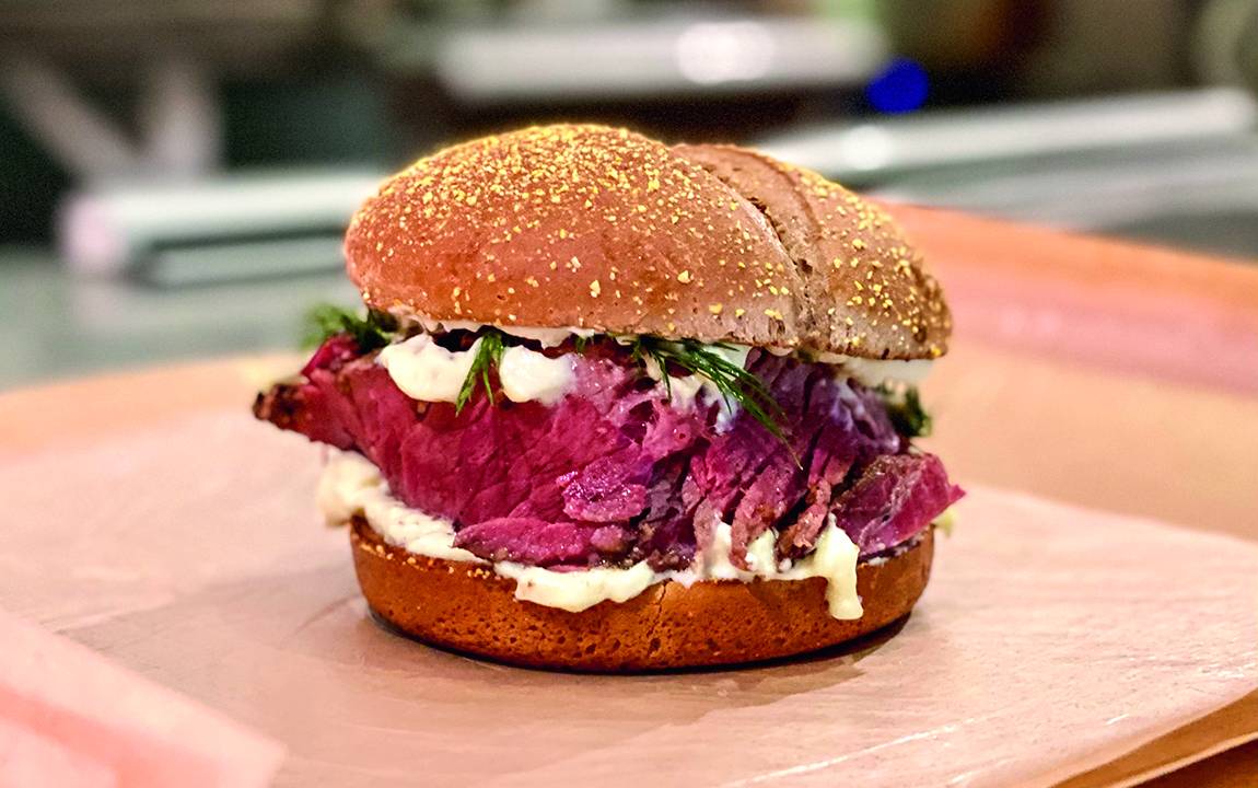 Sanduíche sobre mesa de madeira no pão de hambúrguer escuro, com pastrami arroxeado e maionese aparecendo no recheio.