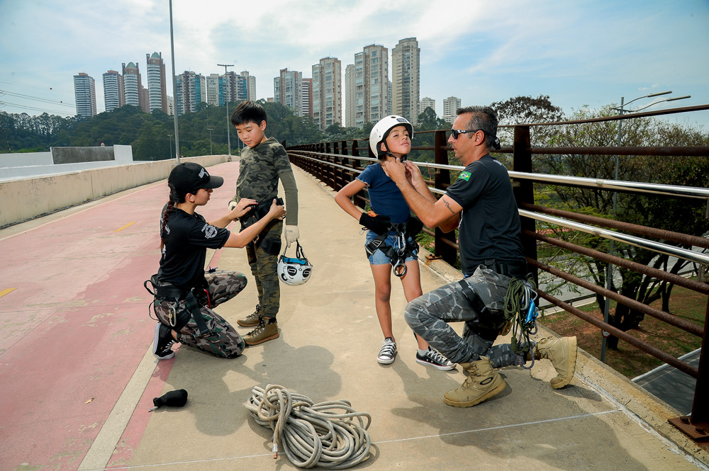 rapel-vertical-infinity-para-criancas-preparacao.jpg em ponte de sp com prédios ao fundo, dois instrutores colocam equipamentos de segurança em duas crianças, para que possam realizar rapel