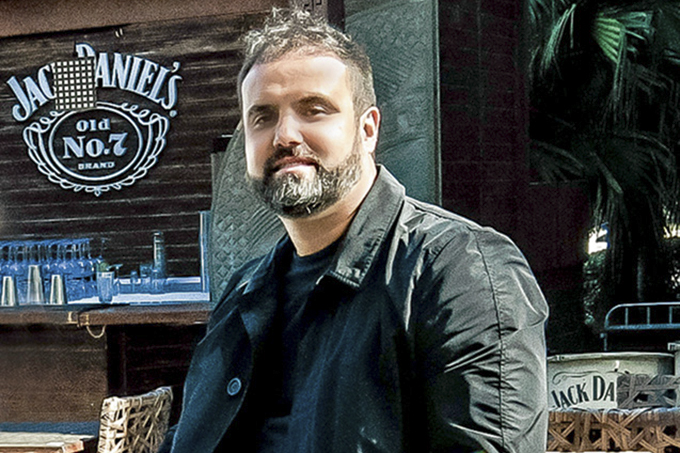 A imagem mostra César Ferreira, sentado em um banquinho, em frente ao seu bar.