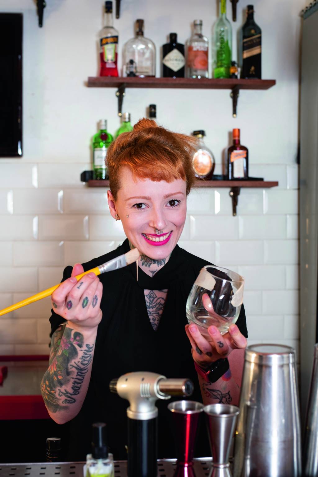 bartender Stephanie – Espaço13 A bartender Stephanie Marinkovic, de cabelo ruivo e braços tatuados, pintando o lado de fora de um copo com ganache de chocolate branco.