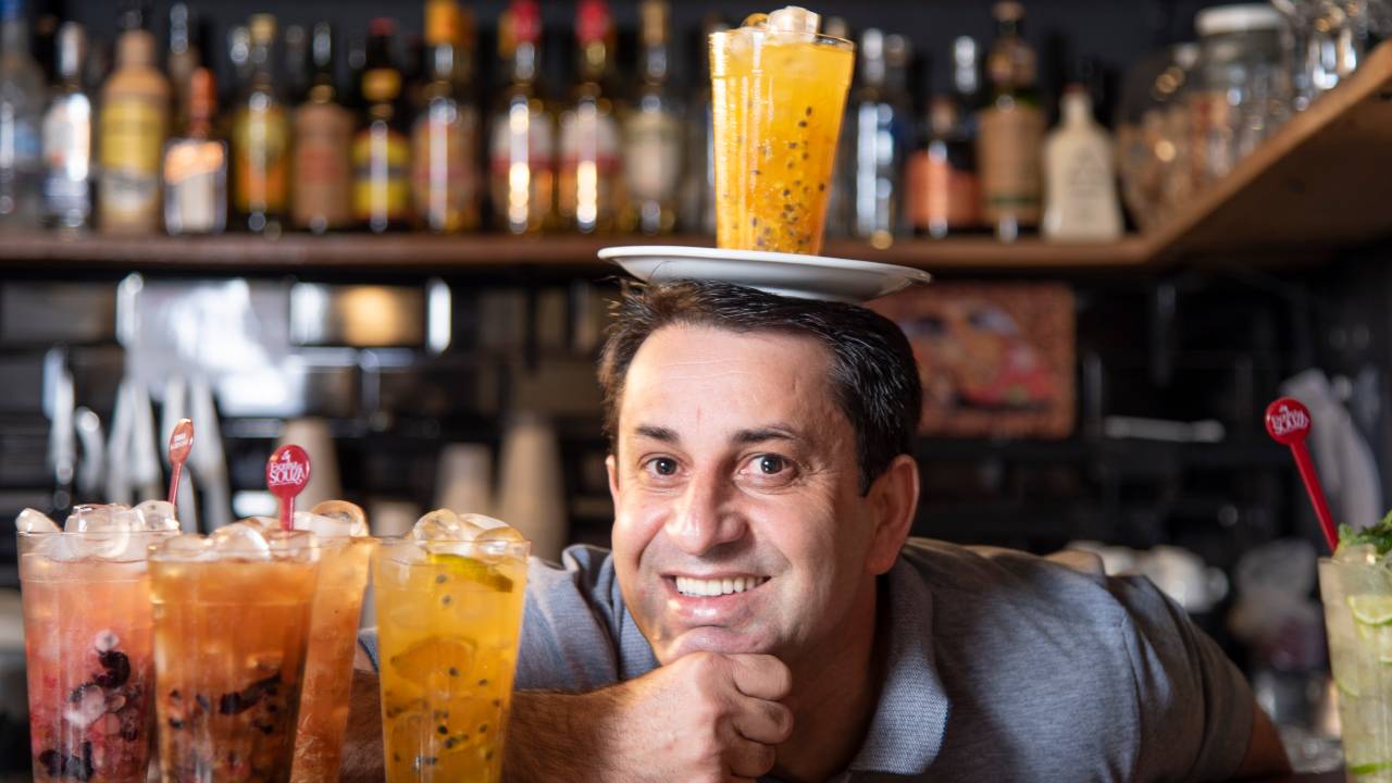 Souza com o rosto apoiado sobre mão cerrada em cima de balcão de madeira do Esquina do Souza ao lado de copos de caipirinha com um prato com uma caipirinha de maracujá na cabeça.