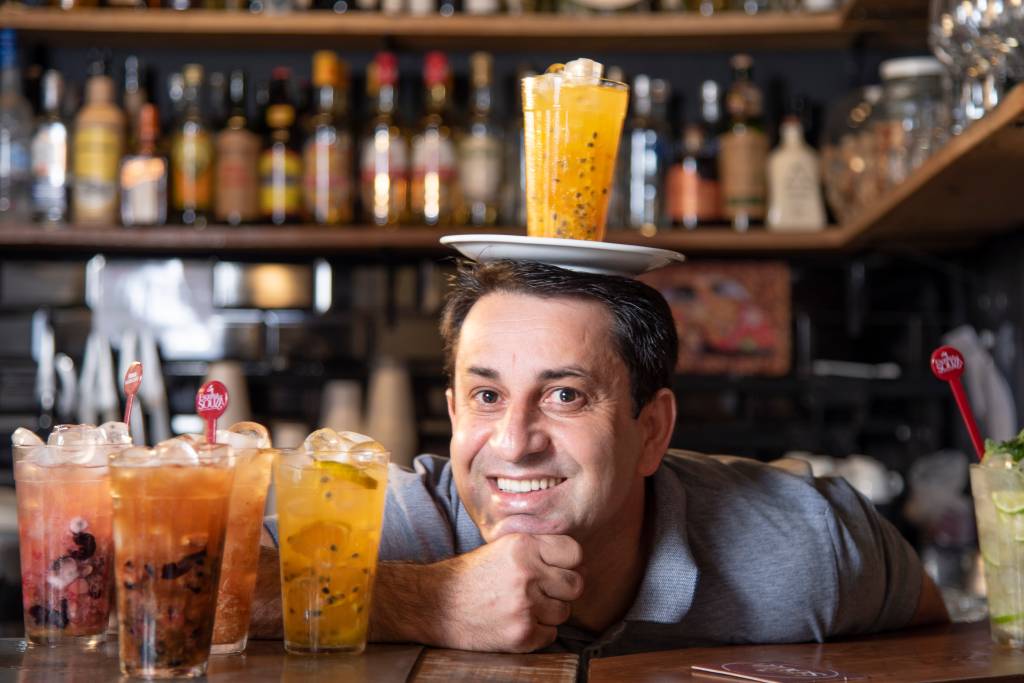 Souza com o rosto apoiado sobre mão cerrada em cima de balcão de madeira do Esquina do Souza ao lado de copos de caipirinha com um prato com uma caipirinha de maracujá na cabeça.