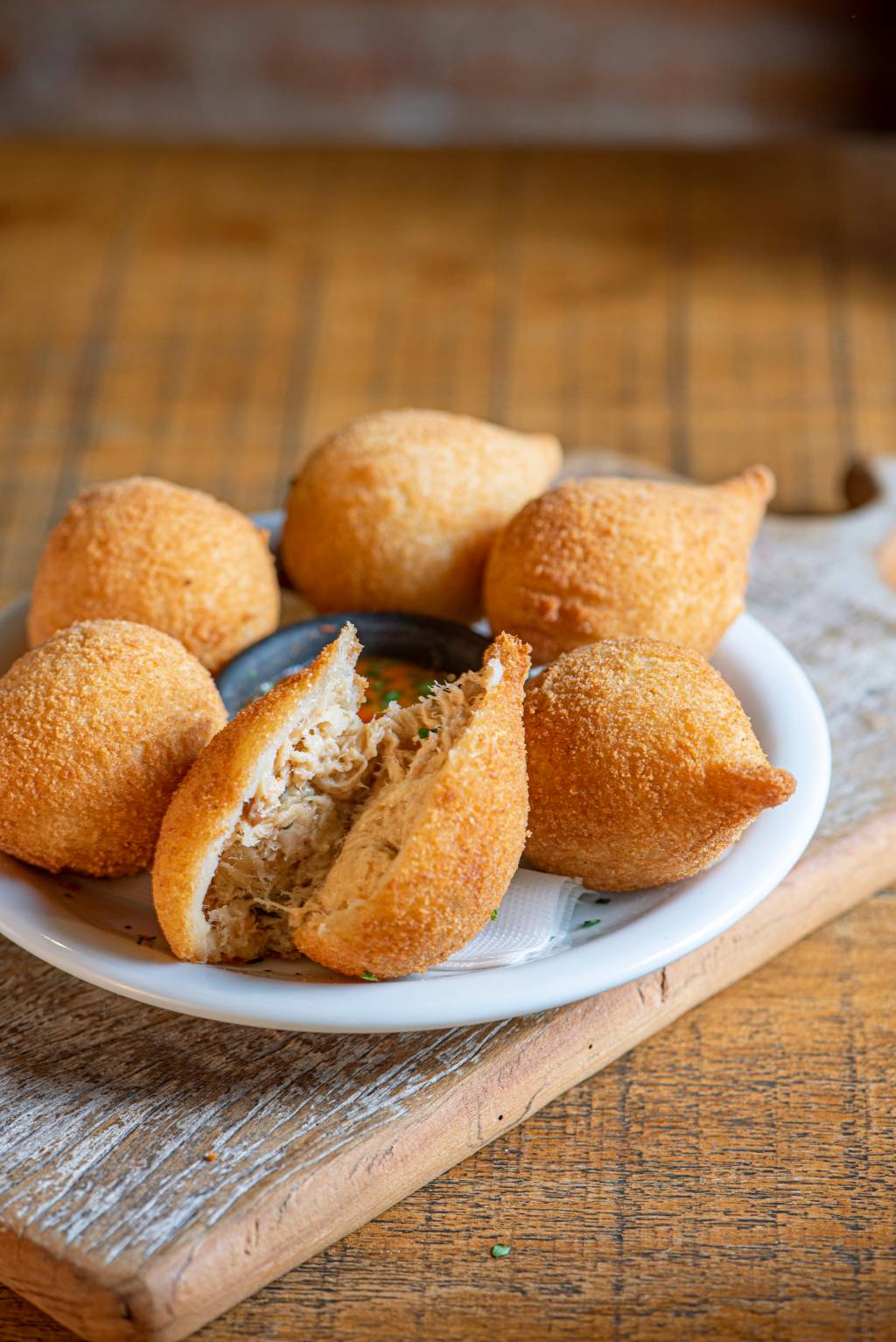 Porção de seis coxinhas sobre prato de louça branca envolta de potinho de molho de pimenta com uma delas, ao centro cortada ao meio com o recheio de frango à mostra.