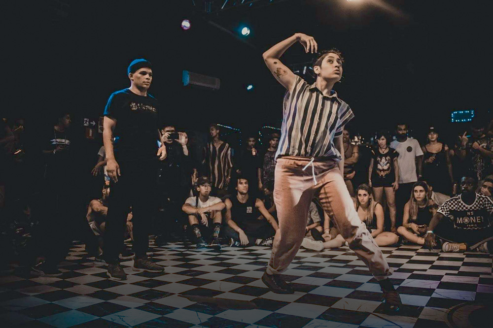 A imagem mostra Giovanna em uma pista de dança, cercada por vários espectadores. Ela está em movimento, com o braço direito levantado e há outra pessoa atrás, em pé, observando.