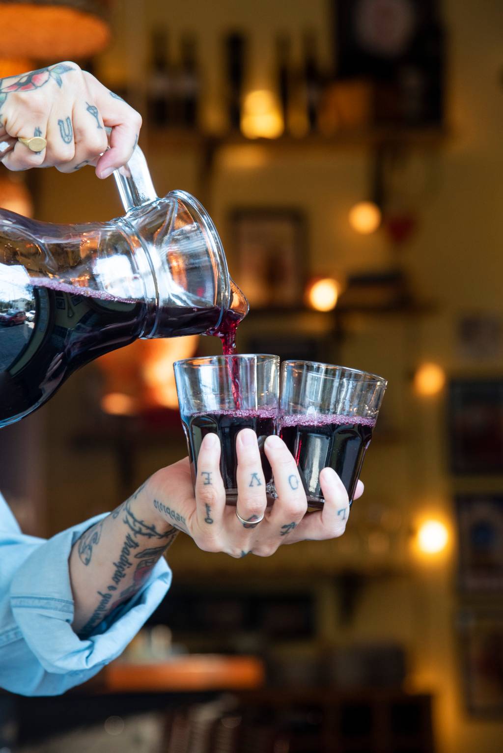 Comer & Beber 2021 – Huevos de Oro Ao centro, mão tatuada segurando dois copos de vidro sendo servidos de vinho em jarra à esquerda.