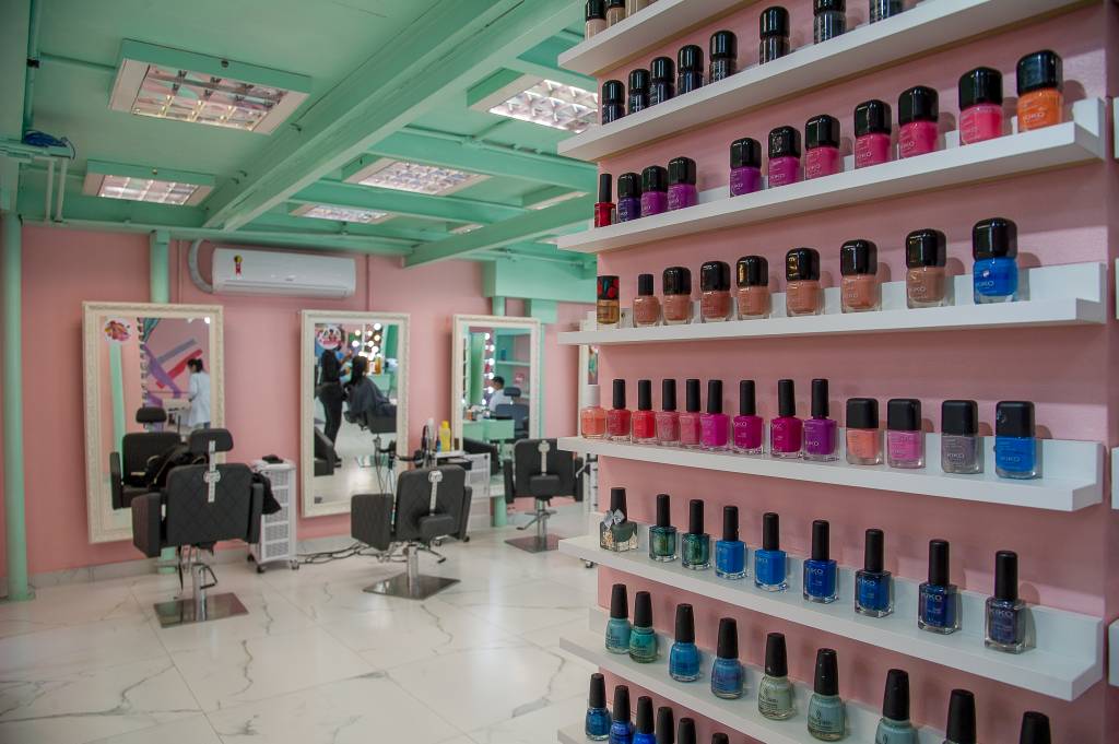 Decoração da barbearia I Love Me exibe pilar cheio de esmaltes e cadeiras ao fundo. Paredes nas cores verde claro e rosa.