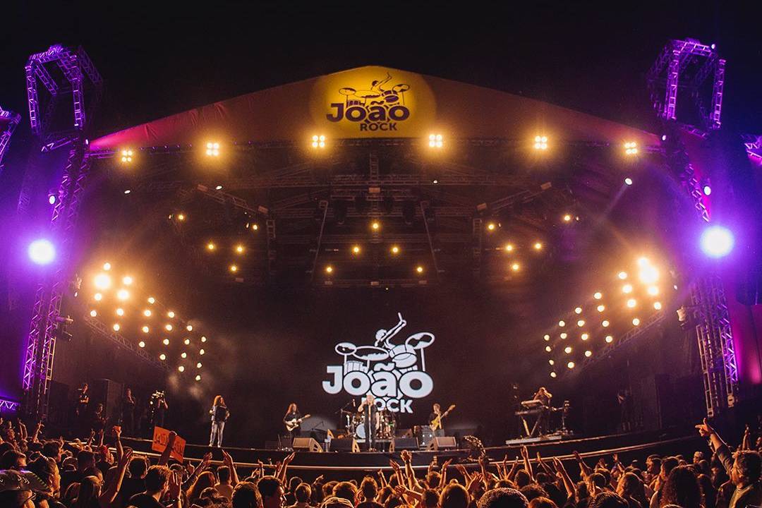 A imagem mostra um palco com muitas pessoas na pista em frente dele. Há uma banda tocando e, no fundo, um logo escrito João Rock