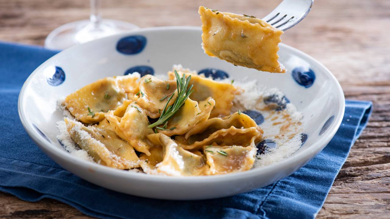 Prato fundo de louça branca com bolinhas azuis com agnollit del plin e garfo levantando um quadrado de agnolotti.