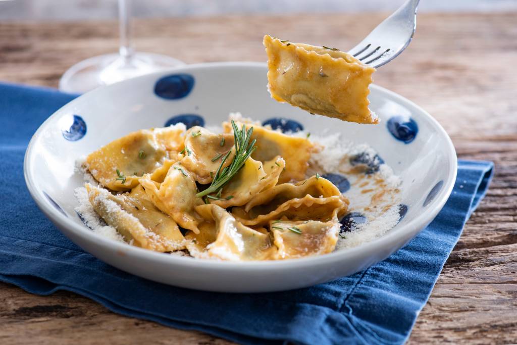 Prato fundo de louça branca com bolinhas azuis com agnollit del plin e garfo levantando um quadrado de agnolotti.