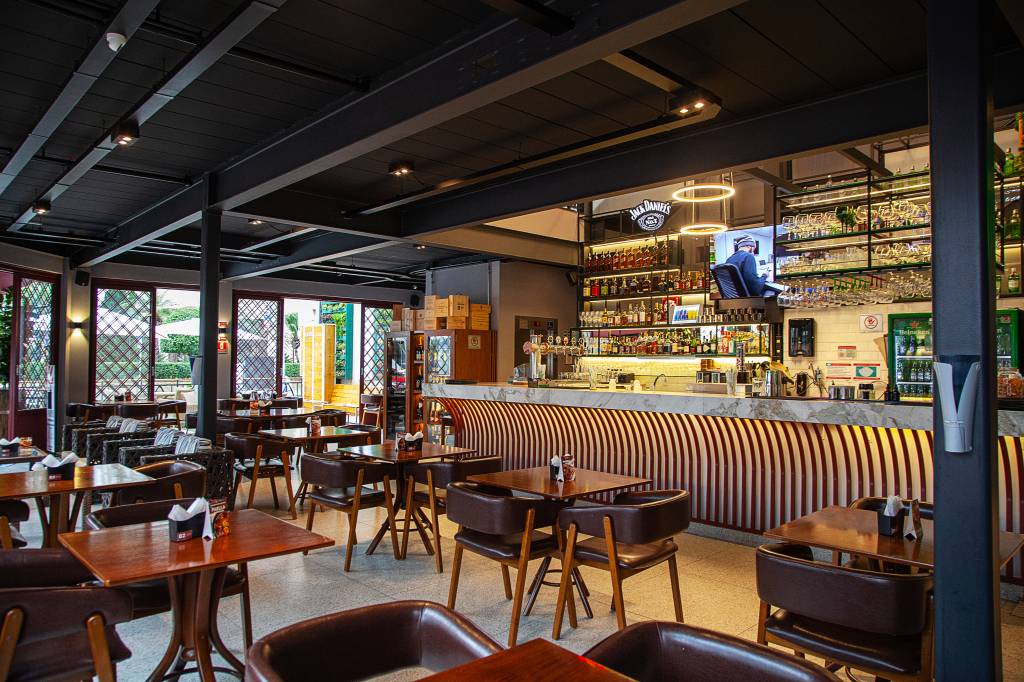 Achados ELO – Martin Bar Salão do Martin Bar com balcão e prateleiras de bebidas à direita e mesas de madeira com cadeiras forradas por couro espalhadas.