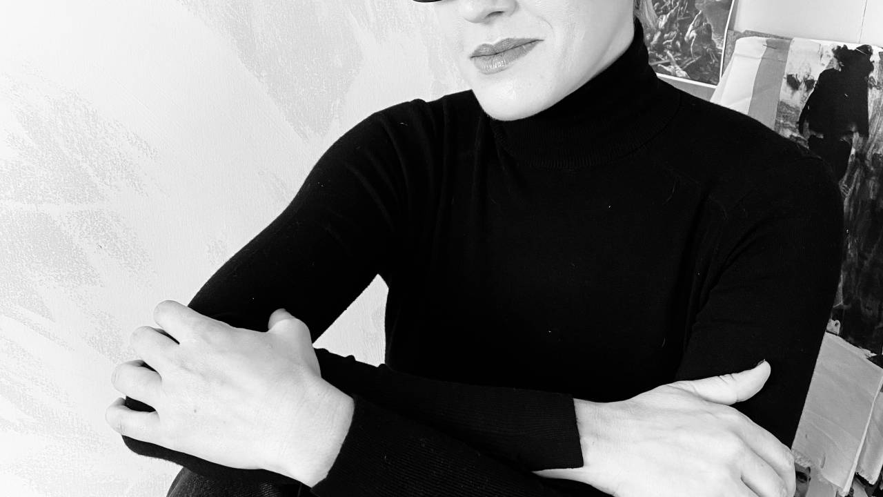 Olivia Merquior posa de braços cruzados em foto preto e branco. Veste suéter preto e usa óculos de armação grossa e quadrada. Tem cabelos curtos.