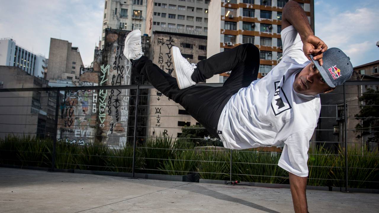 Pelezinho se ergue do chão com uma mão só, com as pernas na horizontal, encarando a câmera. Prédios aparecem ao fundo. Ele veste calça preta, boné e camisa branca.