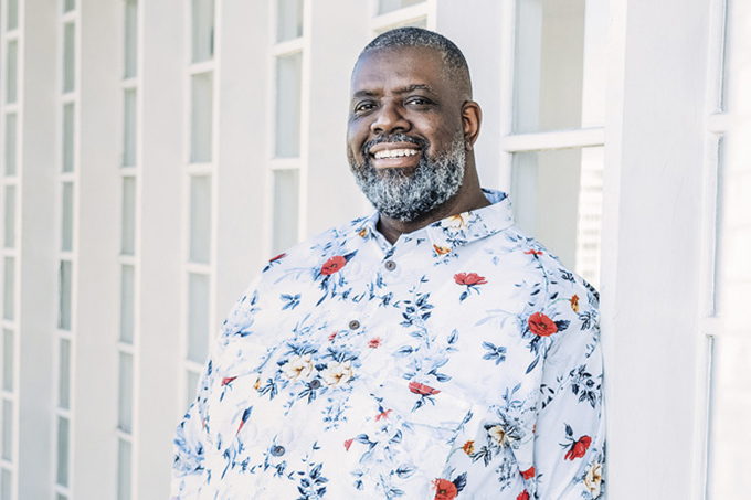 A imagem mostra Péricles, sorridente, com uma camisa florida em uma parede branca olhando para a câmera.