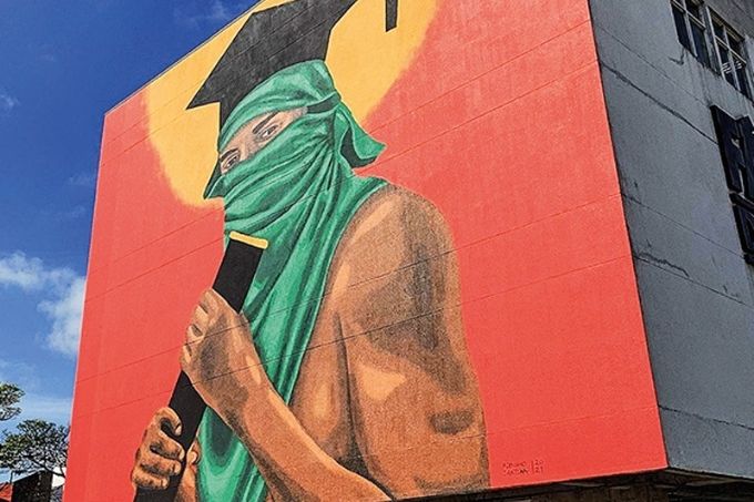 pintura, estampada na parede do prédio da fábrica de cultura de diadema, de homem negro sem camisa com rosto coberto, um chapéu de formando e um tubo de diploma na mão