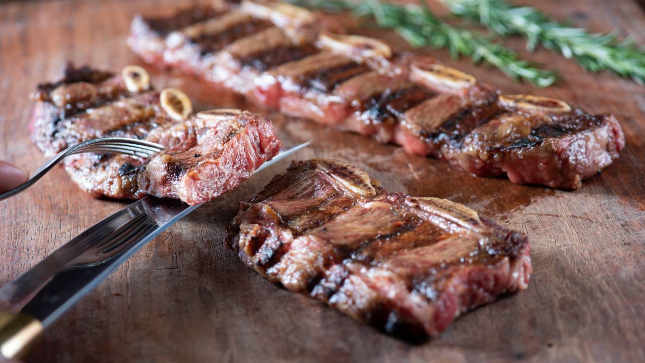 Duas fatias de bife de tira sobre tábua de madeira com faca e garfo segurando um pedaço da carne.