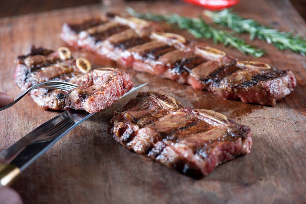 Duas fatias de bife de tira sobre tábua de madeira com faca e garfo segurando um pedaço da carne.