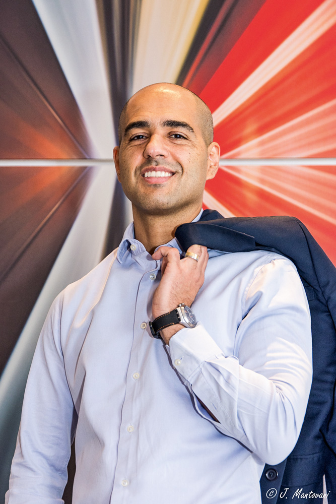 Wladimir Chinchio Um homem de camisa social azul e careca posa sorrindo para a foto. Ele segura um casaco com a mão apoiado nos ombros