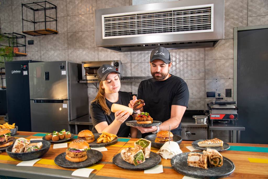 O casal de chefs Giovanna Perrone e Rodrigo Aguiar finalizando sanduíche juntos em cozinha aberta da Zoe Sandwich Shop com vários pratos com sandubas em frente à eles em balcão de madeira.