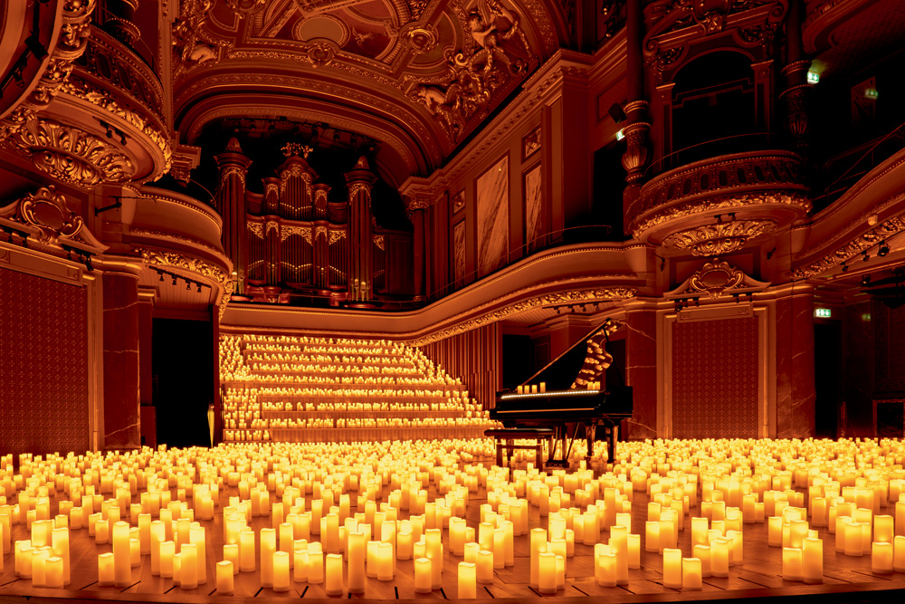 candlelelight experience A foto mostra um grande espaço cheio de velas no chão e em uma escadaria larga em frente. Há um piado de calda, com algumas velas em cima no centro do salão.